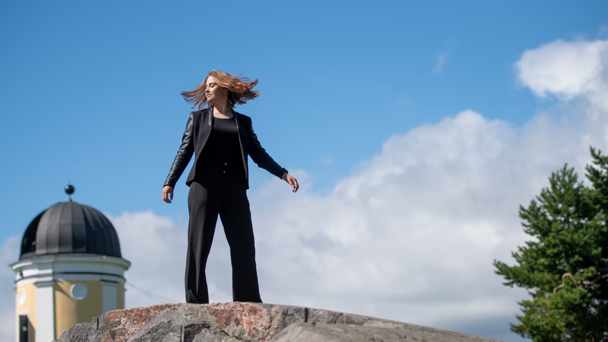 Young woman posing confidently on a rock in a Helsinki park, with a clear blue sky and European architecture in the background.